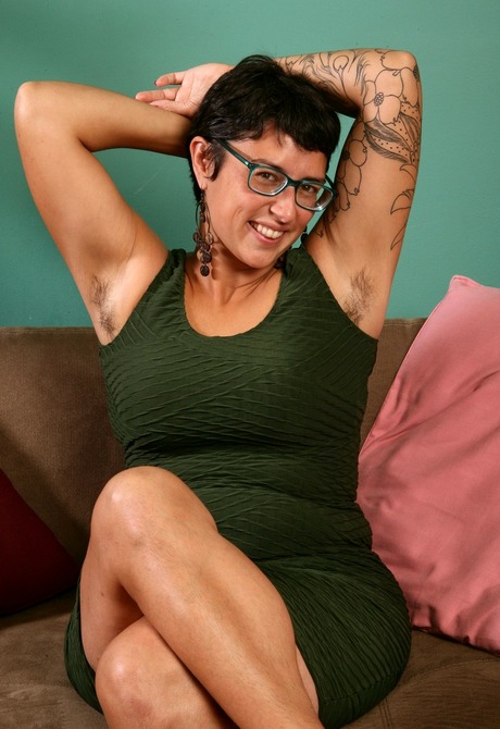 Tattooed brunette Cora teases her bushy beaver on the sofa